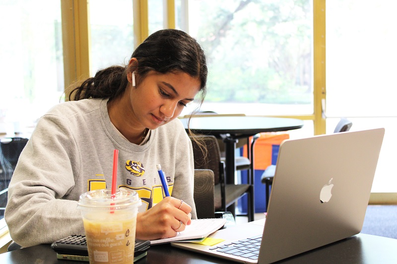 a student takes notes at a desk in the library. A laptop and iced coffee are on the desk in front of her.