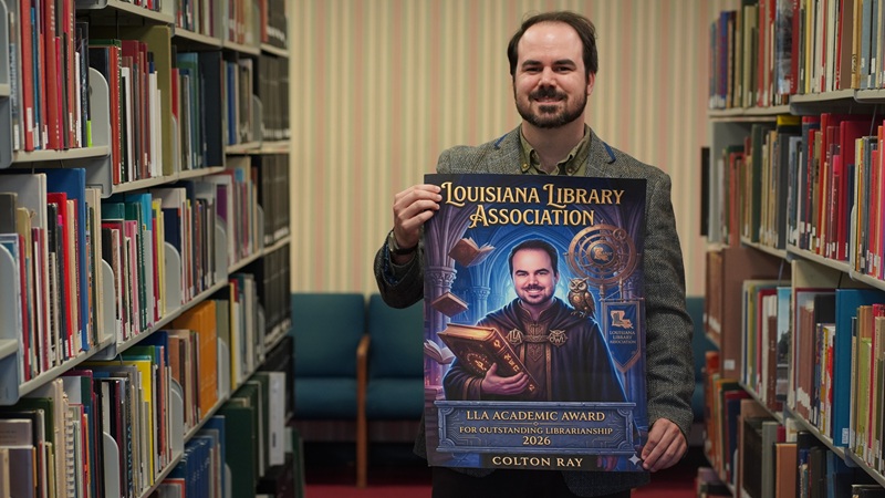 A man is standing between two rows of bookcases in a library setting. He holds up a poster that reads Louisiana Library Association. The image on the poster shows him in a wizard's robe and holding a large book with an owl on his shoulder, reminiscent of Harry Potter.