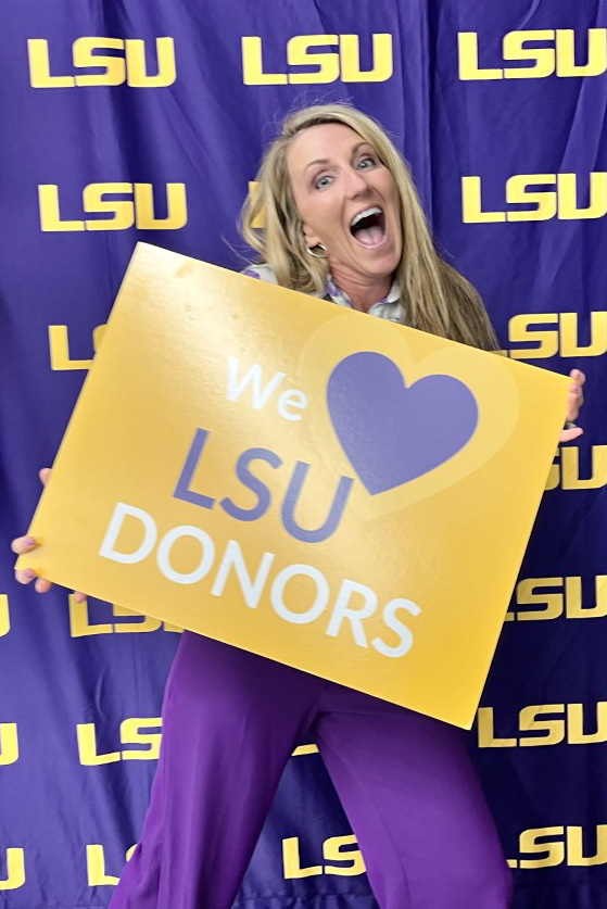Amy Bieger smiles and holds a sign reading We Love LSU Donors