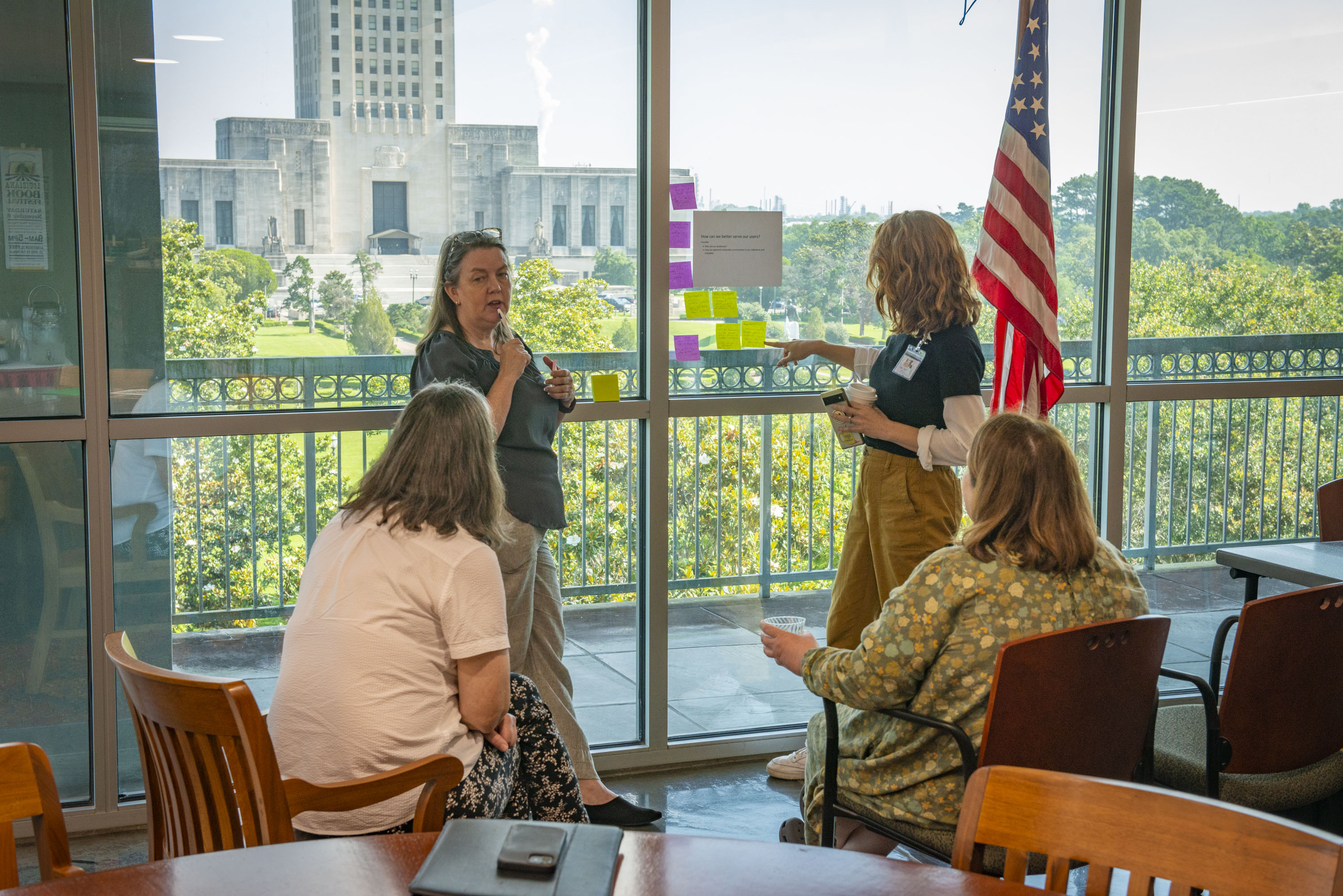 Four women speak and gesture to sticky notes on a window. Louisiana's Capitol Building can be seen through the window.